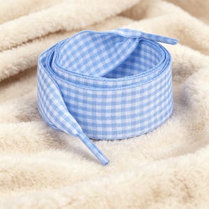 May include: A flat shoelace featuring a light blue and white gingham check pattern. The shoelace is coiled into a circular shape and includes matching light blue and white aglets at both ends, designed for securing footwear with a woven fabric texture.