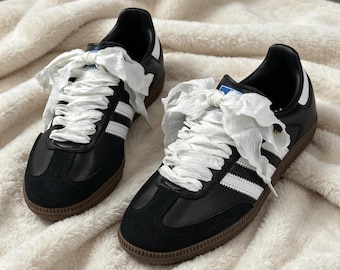 May include: A pair of black sneakers with white leather stripes, black suede toe overlays, and gum rubber soles. The shoes feature wide, white, crinkled ribbon laces tied in large bows at the top, with a blue and white logo patch on the tongue.
