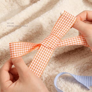May include: An orange and white gingham ribbon tied into a bow, featuring the text SECURE STRONG AND DURABLE printed on the fabric. A separate blue and white gingham ribbon is visible at the bottom, resting against a textured cream fabric.