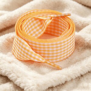 May include: A coiled flat shoelace featuring a vibrant orange and white gingham check pattern. The shoelace is constructed from a woven fabric and includes matching orange and white checkered plastic aglets at both ends of the lace.