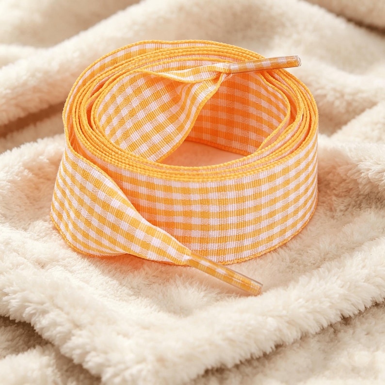 May include: A coiled flat shoelace featuring a vibrant orange and white gingham check pattern. The shoelace is constructed from a woven fabric and includes matching orange and white checkered plastic aglets at both ends of the lace.