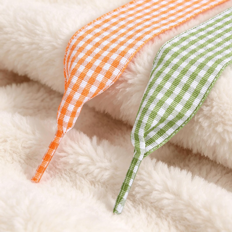 May include: Two flat shoelaces featuring a gingham check pattern, one in bright orange and white and the other in grass green and white. The laces have reinforced plastic aglets at the ends and are resting on a soft, cream-colored fleece fabric.