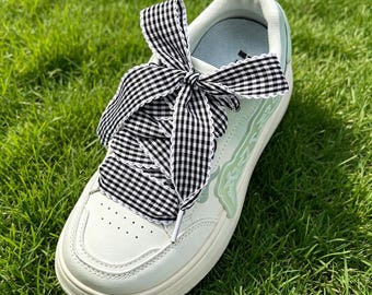 May include: A white sneaker featuring a light green side panel with a decorative design and wide black and white gingham patterned ribbon laces tied in a bow. The shoe has a thick white sole and a small pull tab at the heel collar.