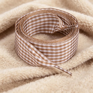May include: A coiled fabric shoelace featuring a brown and white gingham check pattern. The shoelace has a smooth, woven texture and includes clear plastic aglets at the ends, designed for securing footwear with a consistent, repeating grid design.