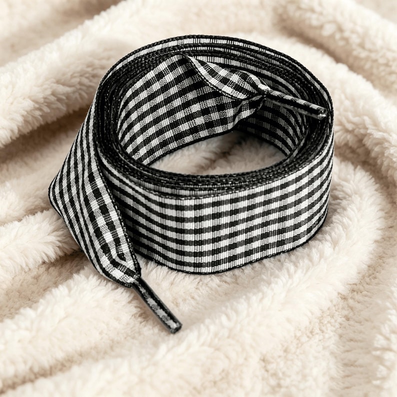 May include: A flat shoelace featuring a black and white gingham check pattern. The shoelace is coiled into a circular shape and includes black plastic aglets at both ends to prevent fraying and facilitate easy threading through eyelets.