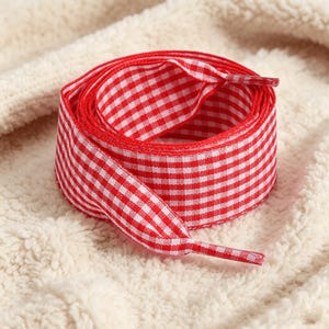 May include: A coiled fabric shoelace featuring a red and white gingham checkered pattern. The shoelace has a flat, woven texture and includes two plastic aglets at the ends, which are also covered in the same red and white checkered design.