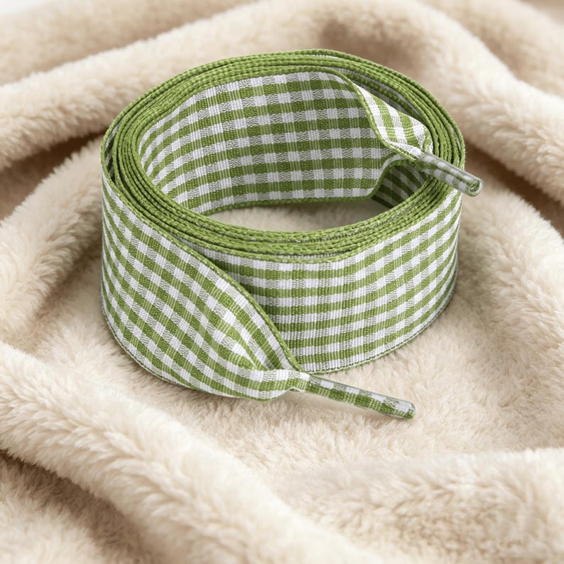 May include: A coiled shoelace featuring a green and white gingham check pattern. The flat fabric lace has clear plastic aglets at both ends, designed for threading through eyelets on footwear to secure the fit of a shoe or sneaker.
