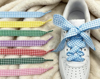 May include: A white sneaker laced with light blue gingham ribbon laces, accompanied by a collection of flat gingham shoelaces in light blue, yellow, green, purple, red, dark green, pink, and black, all featuring a classic checkered pattern.