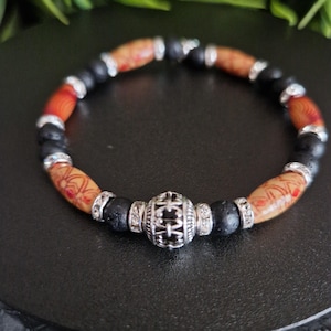 May include: A beaded bracelet featuring alternating black porous lava beads and cylindrical tan beads with red and orange patterns. The design includes silver-toned metal spacers with clear crystals and a central silver-toned filigree bead.
