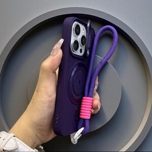 May include: A deep purple smartphone case featuring a circular magnetic charging ring on the back, a camera lens cover, and a purple braided wrist strap accented with a bright pink ribbed silicone connector and a white plastic attachment clip.
