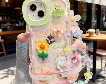 以下が含まれることがあります： ライトグリーンのカメラフレームが付いた透明なスマートフォンケース。3Dのフラワーとカエルのチャーム、「Good Luck」と「Tulip」の文字、そしてカラフルな花のチャームが付いたビーズストラップがぶら下がっています。ケースの下部にはピンクの縁取りがあります。