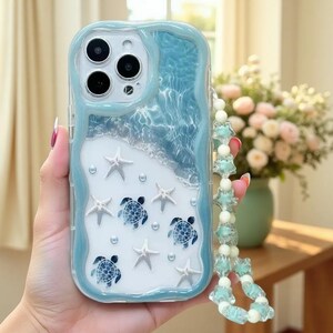 May include: A light blue and white phone case featuring a wavy border, a water-like texture at the top, and a sandy beach design at the bottom. The case is decorated with white starfish, blue sea turtle graphics, and small pearls, with a beaded strap.