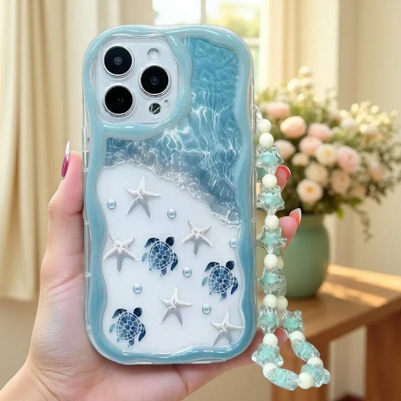 May include: A light blue and white phone case featuring a wavy border, a water-like texture at the top, and a sandy beach design at the bottom. The case is decorated with white starfish, blue sea turtle graphics, and small pearls, with a beaded strap.