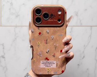 以下が含まれることがあります： 赤、黄、青の散らばった花柄が特徴のタンカラーのスマホケース。カメラモジュールはゴールドの縁取りで、Flower Seaという文字が入ったダークブラウンのインサートが付いています。下部の長方形のラベルには、I am willing to go to a world full of flowers and embrace the sea of flowersと書かれています。
