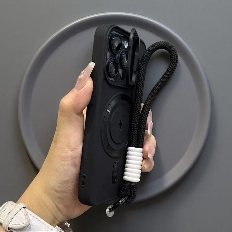 May include: A matte black smartphone case featuring a circular magnetic charging ring on the back, a sliding camera lens cover, and a black braided wrist strap accented with a series of white cylindrical beads attached to the bottom corner.