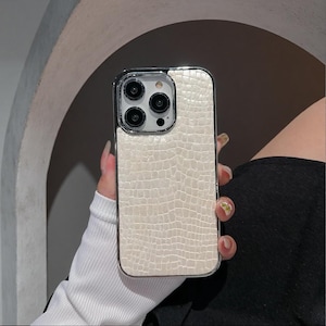 May include: A smartphone case featuring a cream-coloured, textured faux-alligator skin pattern. The case has a glossy, metallic-finished border that frames the back panel and surrounds the camera module, which contains three circular lenses.