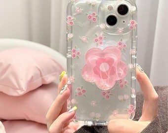 以下が含まれることがあります： 小さなピンクの花と緑の葉が繰り返される模様が描かれた透明なスマートフォンケース。ケースの中央背面には、デバイスをしっかりと保持するための、半透明のピンク色の花びら型グリップが取り付けられています。