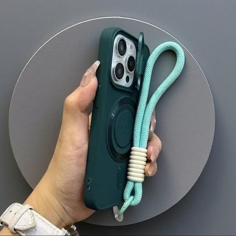 May include: A dark green smartphone case featuring a circular magnetic charging ring on the back, a triple-lens camera cutout with a protective cover, and a light blue braided wrist strap attached with a white coiled accent piece.