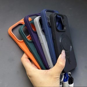 May include: A collection of five smartphone cases in orange, dark green, purple, light gray, and black. Each case features a raised camera cutout, a circular magnetic charging ring on the back, and a bottom attachment point for a lanyard strap.