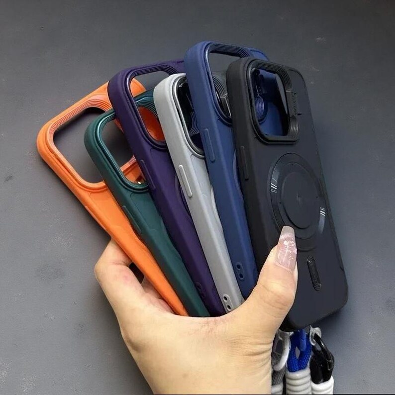 May include: A collection of five smartphone cases in orange, dark green, purple, light gray, and black. Each case features a raised camera cutout, a circular magnetic charging ring on the back, and a bottom attachment point for a lanyard strap.