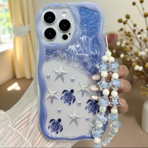 May include: A phone case with a wavy lavender border featuring a blue water-like top section and a white bottom section. The design includes white starfish, blue sea turtle graphics, and small pearls. A beaded strap with star charms is attached.