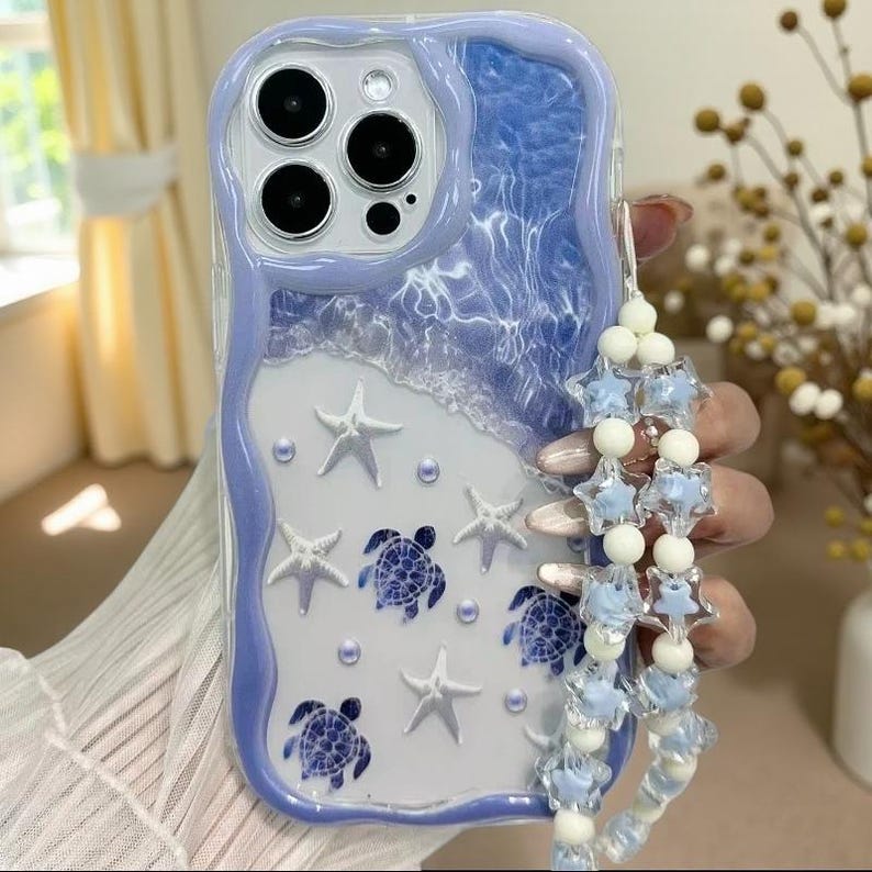 May include: A phone case with a wavy lavender border featuring a blue water-like top section and a white bottom section. The design includes white starfish, blue sea turtle graphics, and small pearls. A beaded strap with star charms is attached.