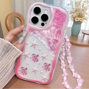 May include: A clear phone case featuring a pink wavy border, a textured pink water-like pattern at the top, and scattered pink starfish and sea turtle embellishments. The case includes a matching pink beaded strap with star-shaped beads.