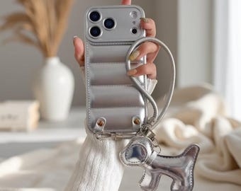 以下が含まれることがあります： キルティング加工が施されたメタリックシルバーのダウンジャケット風スマートフォンケース。手首用ストラップと、取り外し可能なシルバーカラーの犬型チャームが付いています。ケースには3眼カメラ用の切り抜き穴があります。