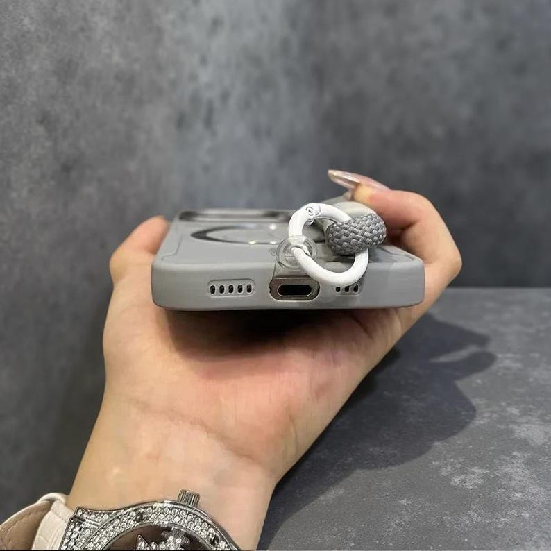 May include: A light gray smartphone case featuring a circular magnetic charging ring on the back and a white plastic ring attachment connected to a short, braided gray cord loop, designed for secure handling and convenient carrying of the device.
