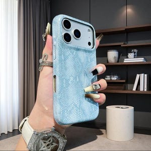Puede incluir: Una funda para smartphone de color azul claro con un patrón texturizado de piel de serpiente. La funda presenta un recorte rectangular para la cámara con tres aberturas circulares para lentes y un sensor de flash.