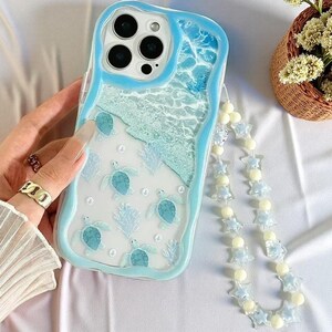May include: A light blue wavy-edged phone case featuring a printed design of sea turtles, coral, and ocean waves. The case includes a matching beaded phone strap made of light blue star-shaped beads and small round white beads.