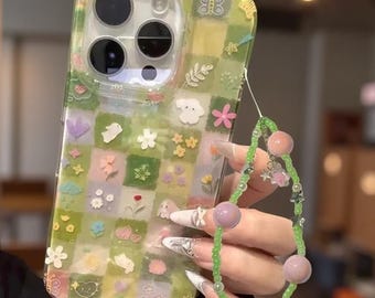 以下が含まれることがあります： 緑と黄色のチェック柄に、小さな花、蝶、動物のモチーフが描かれた透明なスマートフォンケース。小さな緑色のビーズ、大きめの丸いラベンダー色のビーズ、銀色の星のチャームで構成されたビーズストラップが付いています。