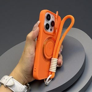 May include: A bright orange smartphone case featuring a circular magnetic charging ring on the back, a sliding camera lens cover, and an attached matching orange woven wrist strap with white decorative beads near the base of the strap.