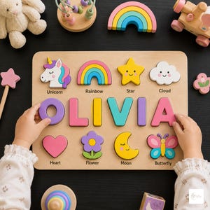 May include: A wooden name puzzle featuring the name OLIVIA in large, colorful block letters. The board includes cutouts for a unicorn, rainbow, star, cloud, heart, flower, moon, and butterfly, each labeled with its corresponding name in black text.