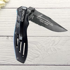 May include: Black folding pocket knife with a partially serrated blade. The knife has the name "DILYNN" engraved on the handle and the text "I'LL ALWAYS BE WITH YOU" on the blade. The knife is open and displayed on a white wooden surface.