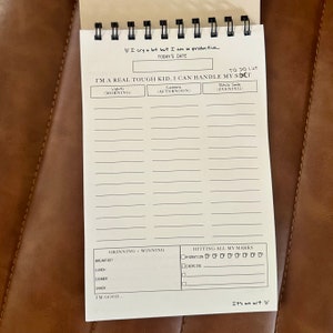 May include: A white planner with a black spiral binding. The planner has a black and white design with the text "I'm a real tough kid, I can handle my s**t" and "Grinning = Winning" and "Hitting all my marks".