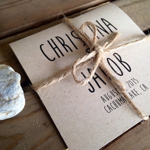 May include: A rustic wedding invitation with twine tied around it. The invitation is printed on brown paper with black text that reads "Christina & Jacob August 1, 2015 Cachuma Lake, CA".