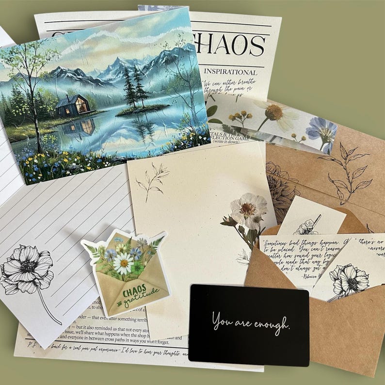 Puede incluir: Un set de papeler&iacute;a que incluye una impresi&oacute;n de un paisaje con una caba&ntilde;a junto a un lago, papel rayado con bocetos florales, una pegatina de un sobre con flores silvestres, una tarjeta negra con el texto You are enough y sobres de papel kraft.