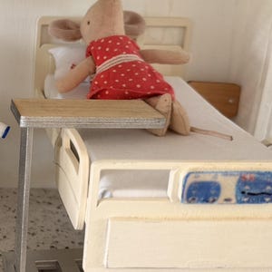 以下が含まれることがあります： 白い水玉模様の赤いドレスを着たベージュの布製ネズミのミニチュア玩具が、明るい木目調のオーバーベッドテーブルが付いた白い病院のベッドに横たわっています。ベッドフレームはクリーム色で、足元には青と白のコントロールパネルが付いています。