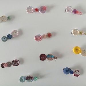 Puede incluir: Una colección de diez pasadores para el cabello dispuestos sobre una superficie, cada uno con una combinación única de botones coloridos y dijes en forma de manzana de diversos tamaños, patrones y texturas, incluyendo tonos de rojo, azul, verde, amarillo y rosa.