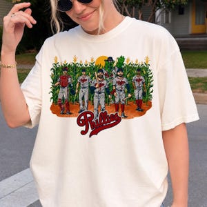 May include: A cream-colored t-shirt featuring a graphic of five baseball players and a green mascot standing in a cornfield. The text Phillies is printed in red script below the illustration. The players wear grey uniforms with red accents.