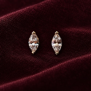May include: A pair of stud earrings featuring marquise-cut clear gemstones set in gold metal. Each stone is secured by two small gold prongs at the top and bottom points, creating a pointed, elongated shape that catches the light.