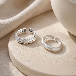 Op de afbeelding: Twee zilverkleurige ringen met een open ontwerp. De ene ring heeft een constructie met meerdere banden en een gedraaide touwtextuur, terwijl de andere ring een glad, effen oppervlak heeft. Beide ringen zijn gemaakt van metaal met een gepolijste afwerking.