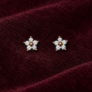 May include: A pair of gold stud earrings shaped like five-petaled flowers. Each earring features five clear, round-cut stones arranged as petals around a central gold bead. The earrings are set against a deep burgundy, textured fabric background.