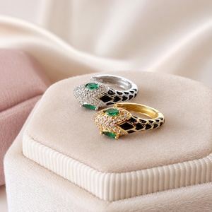 May include: Two hoop earrings shaped like snakes, one in silver tone and one in gold tone. Each earring features a head encrusted with clear pavé stones, two marquise-cut green stones for eyes, and a black enamel scale pattern along the hoop body.