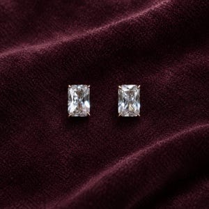 May include: A pair of stud earrings featuring rectangular radiant-cut clear stones set in a metallic frame. The stones have a faceted surface that reflects light, and each is secured by four small prongs at the corners of the rectangular setting.