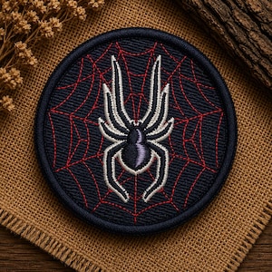 May include: A circular embroidered patch featuring a dark navy blue background with a red stitched spider web pattern. Centred on the patch is a stylised spider with a dark purple body and white outlines on its eight legs, finished with a dark border.