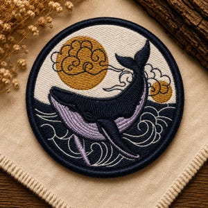 Pode incluir: Um emblema bordado circular apresentando uma baleia azul-escura emergindo de ondas do oceano azul-escuras. O fundo inclui um céu cor de creme com dois sóis ou luas circulares amarelo-dourado e contornos de nuvens estilizados em fio preto.