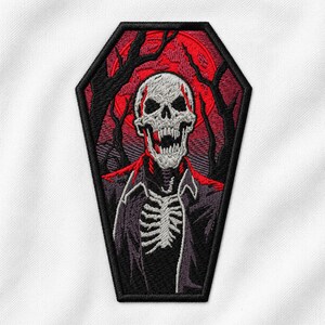 Puede incluir: Un parche bordado con forma de ataúd que muestra una calavera blanca con la boca abierta, una caja torácica y una chaqueta oscura. El fondo representa una luna roja y siluetas de árboles negros, todo dentro de un grueso borde negro.