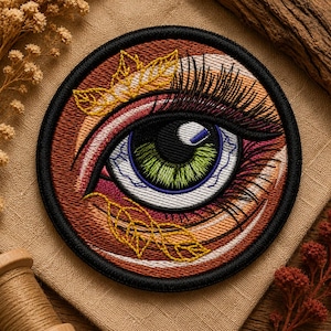 Könnte beinhalten: Ein kreisförmiger bestickter Aufnäher mit einem detaillierten menschlichen Auge mit grüner Iris und schwarzer Pupille. Der umgebende Bereich enthält Braun-, Orange- und Weißtöne, akzentuiert durch gelbe gestickte Blätter und lange schwarze Wimpern.